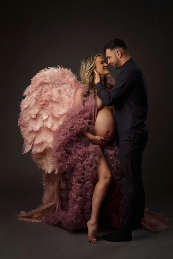 A couple during a maternity photoshoot, with the mother wearing angel wings symbolizing the divine love and protection surrounding their new arrival.