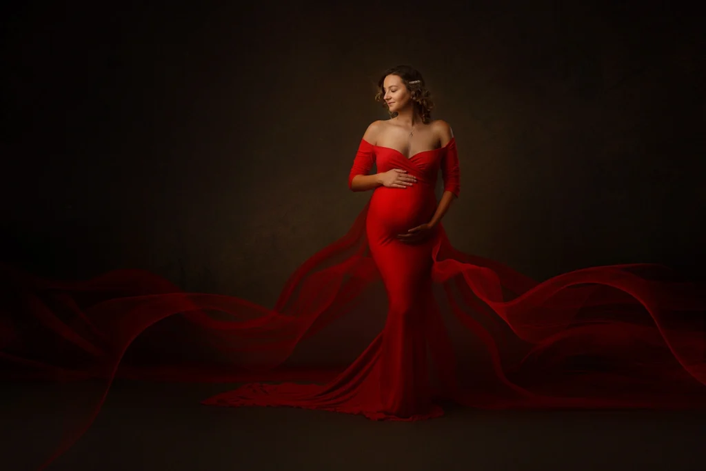 A radiant maternity portrait featuring a woman dressed in a flowing, vibrant red gown. The off-shoulder dress accentuates her elegance and highlights her glowing baby bump. Ethereal, translucent red fabric flows gracefully around her, creating a dynamic and captivating effect. The dark, textured background adds depth and contrast, emphasizing the subject's serene expression and the vibrant red tones of her attire.